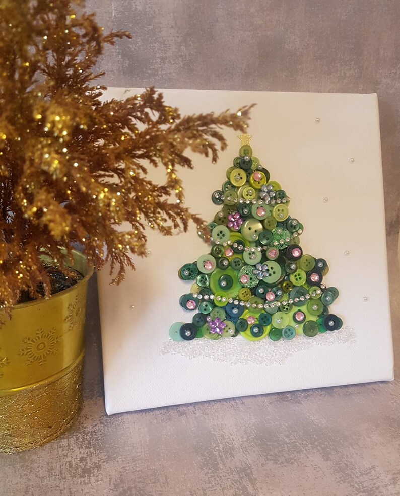 Peut inclure: Une toile blanche avec un sapin de No&euml;l en boutons verts. L'arbre est d&eacute;cor&eacute; de boutons verts, roses et blancs, ainsi que de strass. Le fond est blanc avec quelques points blancs.