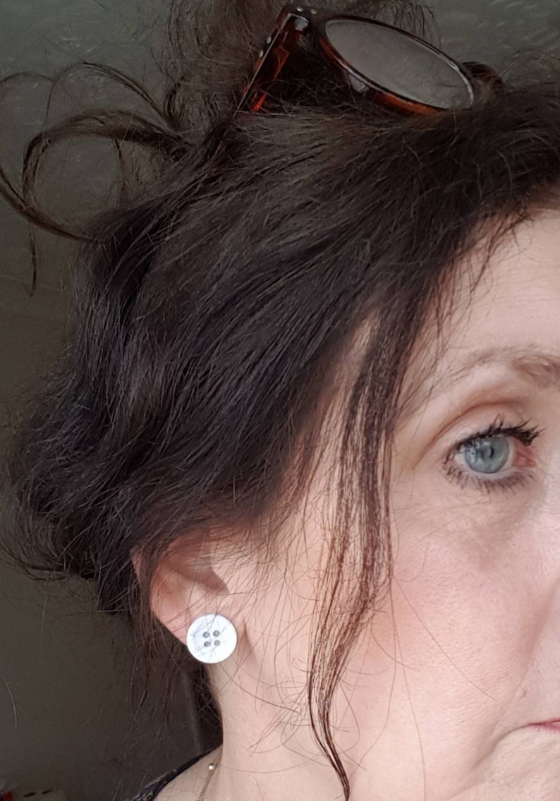 May include: A close-up of a person's ear with a small, round, white button earring. The earring has a four-hole design.