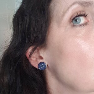 May include: A pair of blue button earrings with white stitching. The earrings are worn in the earlobes of a person with dark hair.