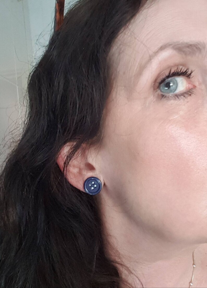 May include: A pair of blue button earrings with white stitching. The earrings are worn in the earlobes of a person with dark hair.