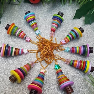 May include: A gold chain necklace with ten colourful button Christmas tree decorations. Each tree is made of stacked buttons in various colours, including red, green, blue, yellow, and purple. The decorations are attached to the chain with clear star-shaped beads.