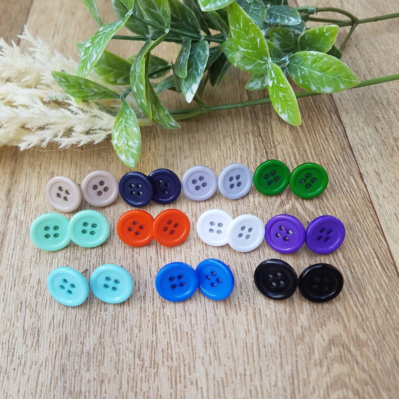 May include: Assortment of colorful button earrings in various colors, including blue, green, purple, red, white, and black. The earrings are arranged in pairs on a wooden surface.