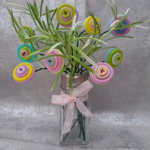 May include: A glass vase with a pink polka dot ribbon around the neck, filled with green stems and colourful button flowers. The buttons are in shades of pink, yellow, blue, and green.