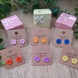 May include: Six pairs of handmade button earrings displayed on brown cards with the text "Handmade with love". The earrings are in various colours, including yellow, orange, purple, and blue.