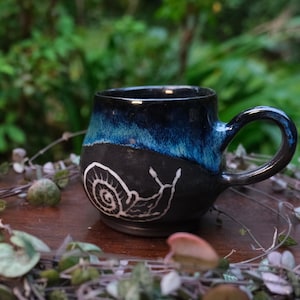 Könnte beinhalten: Eine blaue und schwarze Keramiktasse mit einem weißen Schneckenmotiv. Die Tasse steht auf einem Holztisch mit grünen Ranken.