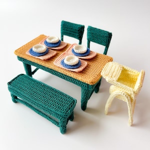 Op de afbeelding: Een gehaakte eettafelset met een groene tafel, stoelen en een bank, en een gele hoge stoel. De tafel is gedekt met oranje placemats en blauwe en witte borden.