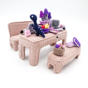 Op de afbeelding: Een roze gehaakte tafel- en bankset met een verscheidenheid aan gehaakte speelgoed en accessoires, waaronder een paars kasteel, een blauwe dinosaurus en een mand met garen.