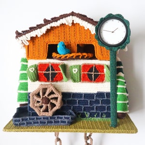 May include: A crocheted wall hanging featuring a small house with a clock, a bird, and a water wheel. The house is orange, green, and white with red and green windows. The clock is green with black hands and a white face. The bird is blue and is perched in a nest. The water wheel is brown and is made of yarn.