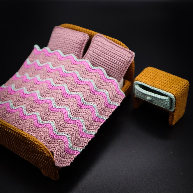 May include: A crocheted dollhouse bed with a pink and white chevron patterned blanket, two pillows, and a matching brown nightstand.