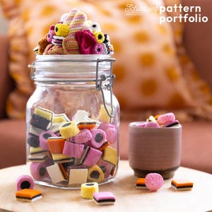 Op de afbeelding: Een glazen pot gevuld met kleurrijke snoepjes en bedekt met gehaakte lekkernijen. De pot staat naast een kleine keramische beker met meer snoepjes. De afbeelding bevat de tekst "Scheepjes pattern portfolio".