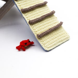 May include: A red crocheted crab toy sits in front of a crocheted play structure with a yellow and brown ladder.