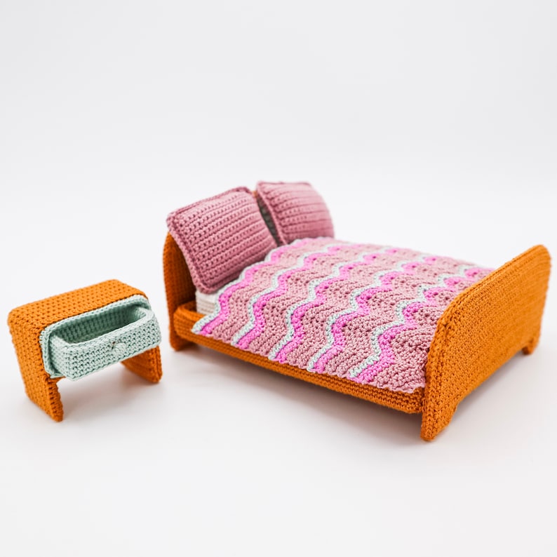 May include: A miniature crocheted bed set with a pink and white chevron patterned blanket, two pink pillows, and a matching orange bedside table with a drawer.