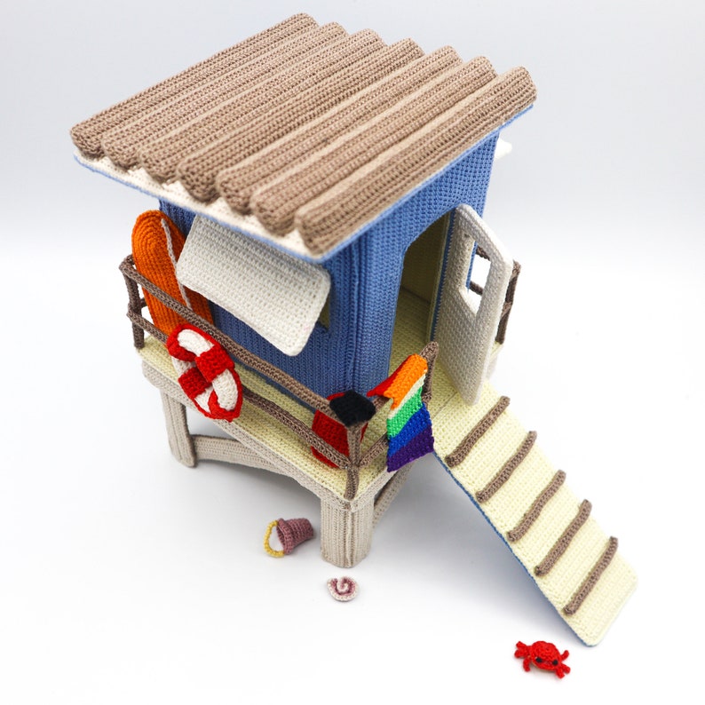 May include: A crocheted beach hut with a brown roof, blue walls, and a white awning. The hut has a small porch with a life preserver, a surfboard, and a rainbow flag. There is a small red crab and a yellow bucket on the ground.