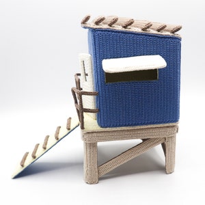 May include: A blue and brown crocheted playhouse with a yellow ladder. The playhouse has a white awning over a window and a brown roof.