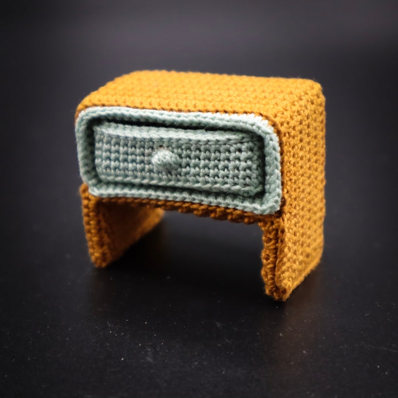 May include: A small crocheted nightstand with a drawer, made of brown and light blue yarn.