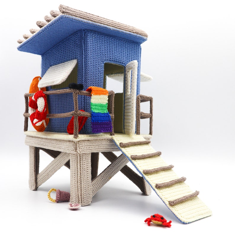 May include: A crocheted lifeguard stand with a rainbow towel hanging inside, a red and white life preserver, and a small red crab on the ground.