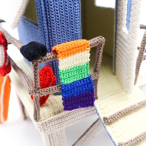 May include: A crocheted beach hut with a rainbow colored towel hanging on the railing. The hut has a white door and a brown ladder.