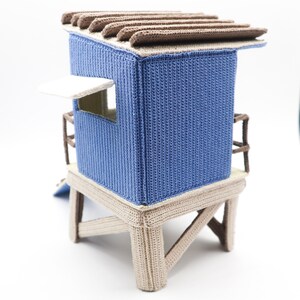 May include: A blue and brown crocheted beach hut toy with a white awning and a ladder. The hut is on a wooden platform with four legs.