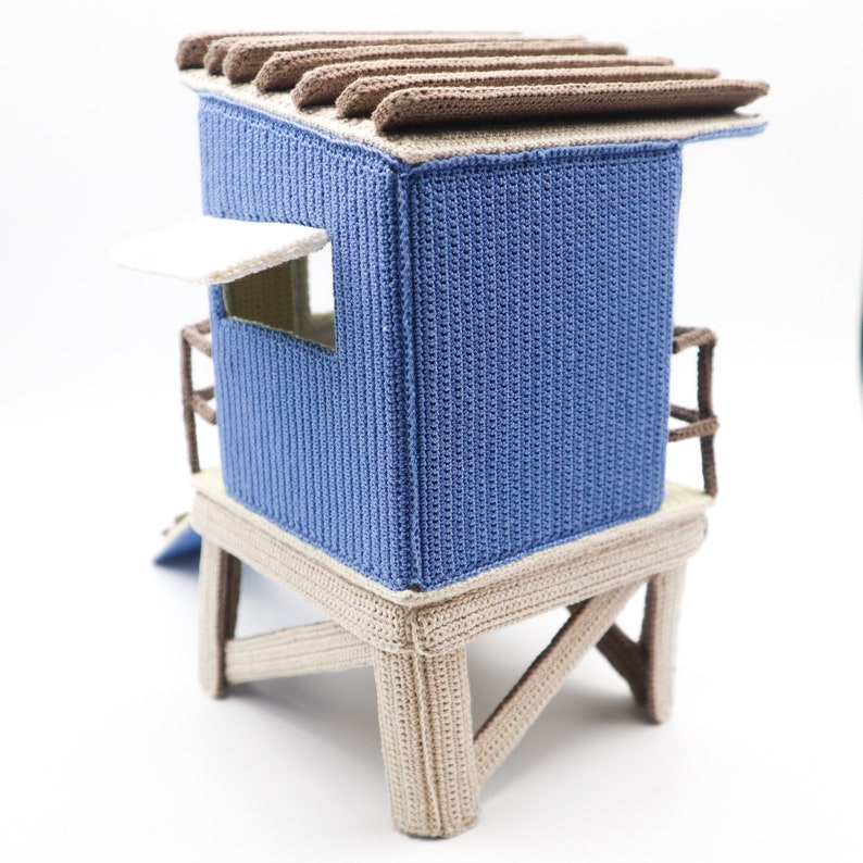 May include: A blue and brown crocheted beach hut toy with a white awning and a ladder. The hut is on a wooden platform with four legs.