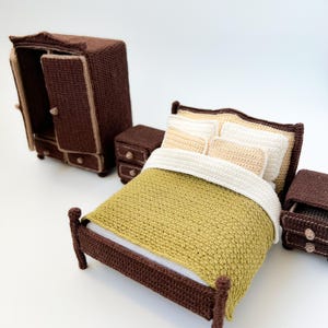 May include: A miniature crochet bedroom set in shades of brown and cream. The set includes a bed with a green blanket and white pillows, a wardrobe, and two nightstands. The wardrobe door is open, revealing the interior.