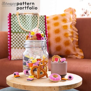 Op de afbeelding: Een houten tafel toont een glazen pot gevuld met kleurrijke gehaakte snoepjes, een pretzel en een klein kopje met meer snoepjes. Een groen-witte geruite draagtas en een geel zonnebloemkussen staan op de achtergrond. De tekst "Scheepjes pattern portfolio" staat bovenaan.