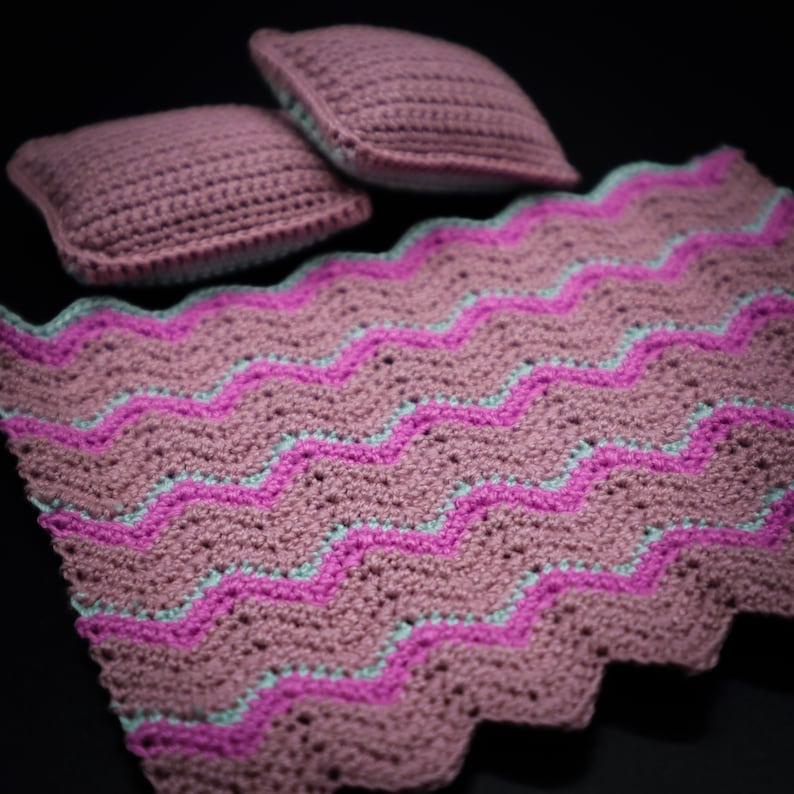 May include: A pink and light blue crocheted blanket with two matching pillows. The blanket has a chevron pattern.