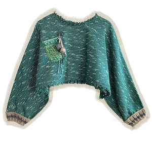 Könnte beinhalten: Ein grüner, gecroppter Pullover mit weißen Flecken und einem ausgefransten Ausschnitt und Saum. Die langen Ärmel haben braun-blau gestreifte Bündchen. Eine kleine Tasche auf der linken Vorderseite ist mit einem grünen Aufnäher und dekorativen hängenden Perlen verziert. Der Pullover hat ein einzigartiges, strukturiertes Aussehen.