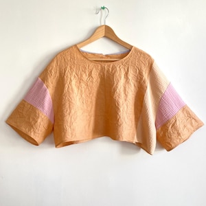 May include: A cropped, short-sleeved top in a textured, peach-colored fabric. The sleeves feature a pink and peach color block design. The top is hanging on a wooden hanger against a white wall.