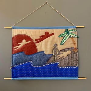 May include: Textile wall hanging featuring a beach scene with figures, a red sun, and a palm tree. The artwork uses various textures and colors, including blue, beige, and green, and is mounted on wooden dowels with a string for hanging.