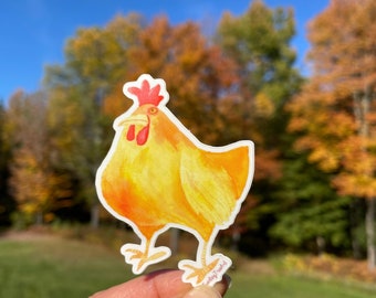 May include: A yellow watercolor chicken sticker with a red comb and wattle. The sticker is held in a hand against a background of fall foliage.