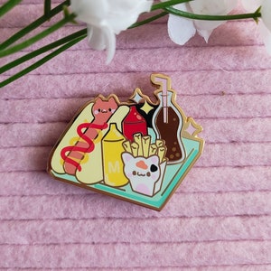 Op de afbeelding: Een gouden emaille pin met een cartoon hotdog, friet, een frisdrank en ketchup. De pin is op een lichtblauwe achtergrond met een gouden rand.