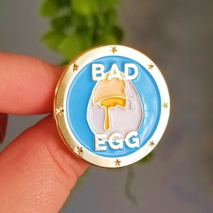 May include: A gold-colored pin with a blue background and white lettering. The pin says "Bad Egg" and features a cartoon image of a cracked egg with yolk dripping out.