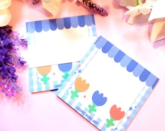 May include: Two blue and white striped notepads with a blue awning and flower designs. The top notepad has a blank space for writing. The bottom notepad has the text "COSMIC BUNS ART" and three colorful flowers.