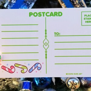 May include: A white postcard with green text that says "POSTCARD" at the top. The postcard has a green line down the center with two green hearts. There are lines for writing a message and an address. The bottom of the postcard has a green candy cane design and the text "@COSMIC BUNS ART".