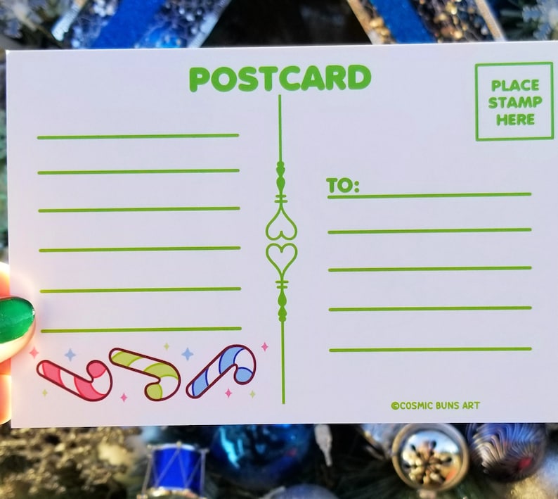May include: A white postcard with green text that says "POSTCARD" at the top. The postcard has a green line down the center with two green hearts. There are lines for writing a message and an address. The bottom of the postcard has a green candy cane design and the text "@COSMIC BUNS ART".