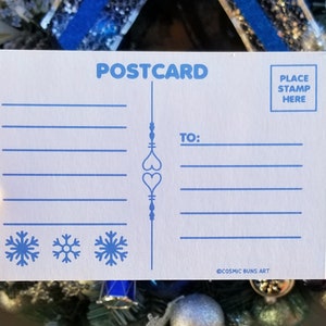 May include: A white postcard with blue text and illustrations. The postcard says "POSTCARD" at the top and "TO:" in the middle. There are three snowflakes at the bottom and the text "COSMIC BUNS ART" at the bottom right. The text "PLACE STAMP HERE" is in a box at the top right.
