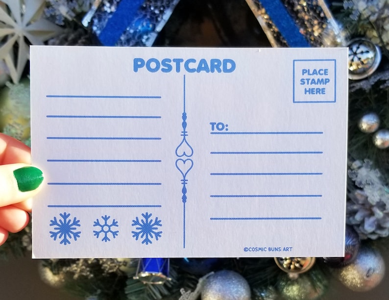 May include: A white postcard with blue text and illustrations. The postcard says "POSTCARD" at the top and "TO:" in the middle. There are three snowflakes at the bottom and the text "COSMIC BUNS ART" at the bottom right. The text "PLACE STAMP HERE" is in a box at the top right.