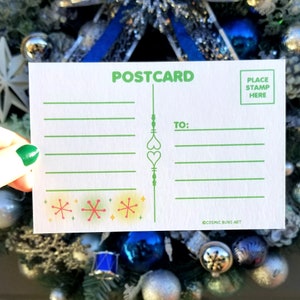 May include: A white postcard with green lines and a green border. The postcard says "POSTCARD" in green letters at the top. There is a space for a stamp in the top right corner with the text "PLACE STAMP HERE". The postcard has a green line down the middle with a heart design. The text "TO:" is written in green letters on the right side of the postcard. The bottom of the postcard has a row of four colorful snowflakes and the text "COSMIC BUNS ART" in small green letters.