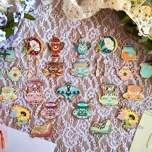 May include: A collection of colorful enamel pins with a tea party theme. The pins include teacups, teapots, spoons, books, and floral designs. The pins are in various colors, including blue, pink, and gold, and are arranged on a lace surface.
