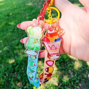 May include: Two acrylic keychains featuring dessert illustrations. One shows a green drink with whipped cream and a blue strap with fruit icons. The other shows a fruit parfait with a cherry on top and a tan strap with various fruit graphics.