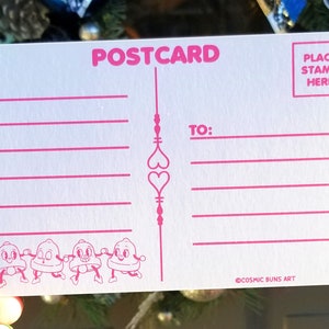 May include: A pink and white postcard with a design of four cartoon bells holding hands. The postcard has lines for writing a message and an area for a stamp. The text "POSTCARD" is printed in pink at the top of the card. The text "PLACE STAMP HERE" is printed in pink in the top right corner. The text "TO:" is printed in pink in the top right corner. The text "COSMIC BUNS ART" is printed in pink at the bottom of the card.
