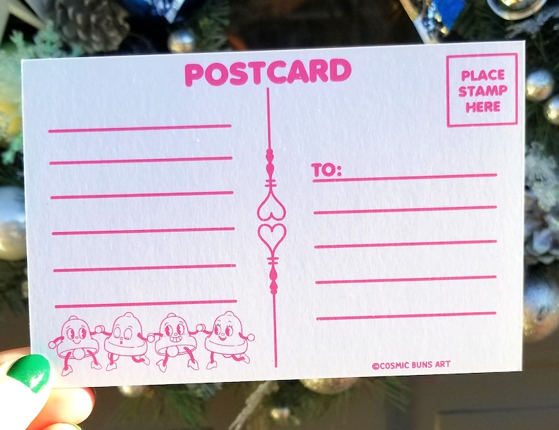 May include: A pink and white postcard with a design of four cartoon bells holding hands. The postcard has lines for writing a message and an area for a stamp. The text "POSTCARD" is printed in pink at the top of the card. The text "PLACE STAMP HERE" is printed in pink in the top right corner. The text "TO:" is printed in pink in the top right corner. The text "COSMIC BUNS ART" is printed in pink at the bottom of the card.