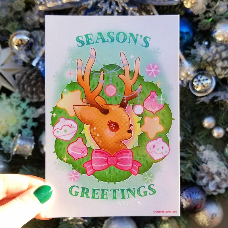 May include: A Christmas card with a green wreath featuring a cute cartoon deer with a pink bow. The wreath is decorated with stars, ornaments, and snowflakes. The card reads "Season's Greetings" and is signed "Cosmic Buns Art".