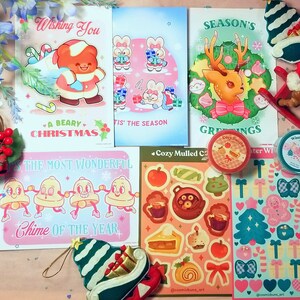 May include: A collection of Christmas-themed stickers and cards featuring illustrations of a bear, reindeer, bells, and a gingerbread house. The stickers are on a pink and blue background with white snowflakes. The cards are on a white background with colourful illustrations.