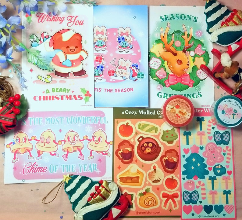 May include: A collection of Christmas-themed stickers and cards featuring illustrations of a bear, reindeer, bells, and a gingerbread house. The stickers are on a pink and blue background with white snowflakes. The cards are on a white background with colourful illustrations.