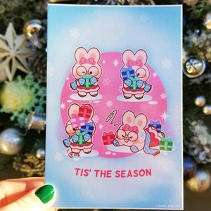 May include: A pink and blue Christmas card with four cartoon bunnies wearing scarves and holding presents. The card has a pink background with snowflakes and the text "Tis' the Season".