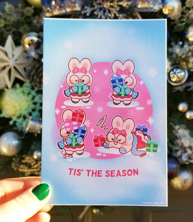May include: A pink and blue Christmas card with four cartoon bunnies wearing scarves and holding presents. The card has a pink background with snowflakes and the text "Tis' the Season".