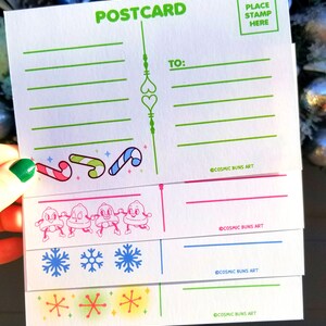 May include: A set of four blank postcards with a winter holiday theme. The postcards feature illustrations of candy canes, snowmen, snowflakes, and stars. The text "POSTCARD" is printed in green at the top of the card. The text "TO:" is printed in green on the right side of the card. The text "COSMIC BUNS ART" is printed in black at the bottom of each card. The text "PLACE STAMP HERE" is printed in a green square at the top right corner of the card.