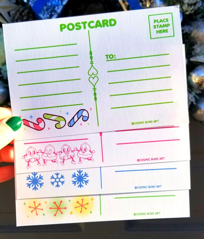 May include: A set of four blank postcards with a winter holiday theme. The postcards feature illustrations of candy canes, snowmen, snowflakes, and stars. The text "POSTCARD" is printed in green at the top of the card. The text "TO:" is printed in green on the right side of the card. The text "COSMIC BUNS ART" is printed in black at the bottom of each card. The text "PLACE STAMP HERE" is printed in a green square at the top right corner of the card.