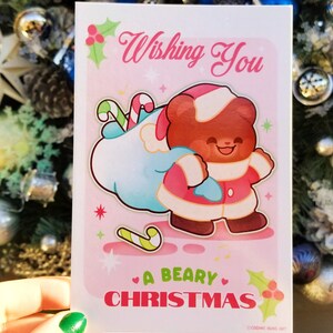 May include: A pink Christmas card with a cartoon bear wearing a Santa hat and carrying a bag of candy canes. The card says "Wishing You A Beary Christmas".